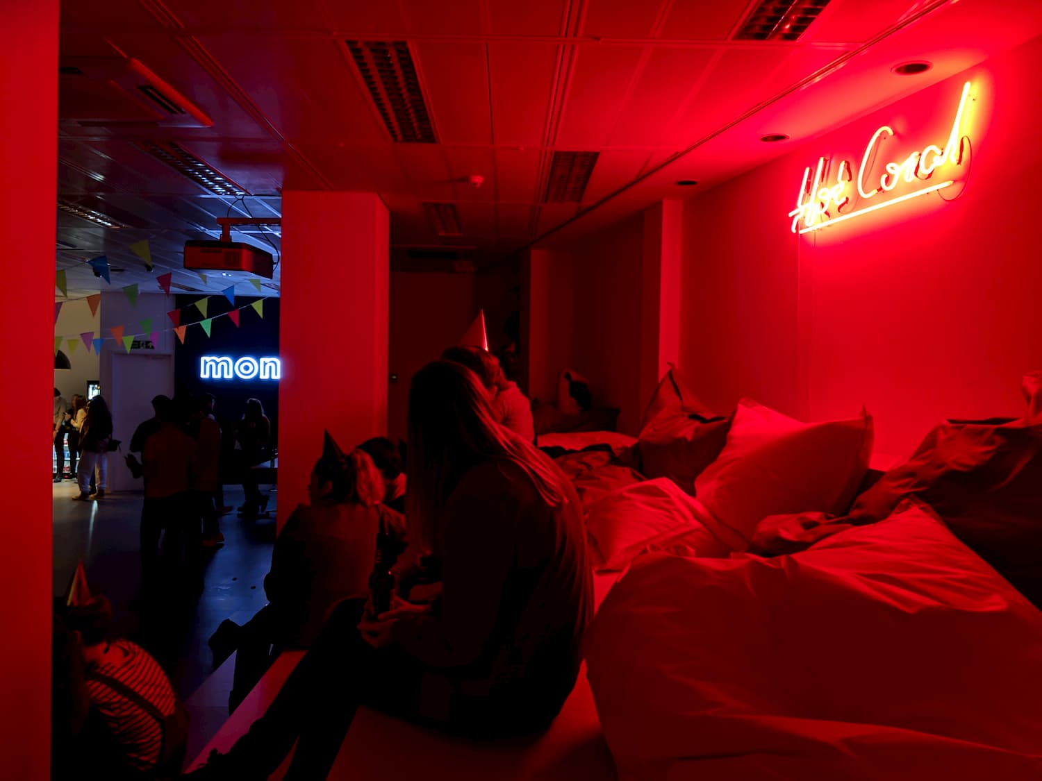 On the right wall a neon sign reads 'Hot Coral' and is projecting a pink glow onto people sat wearing party hats