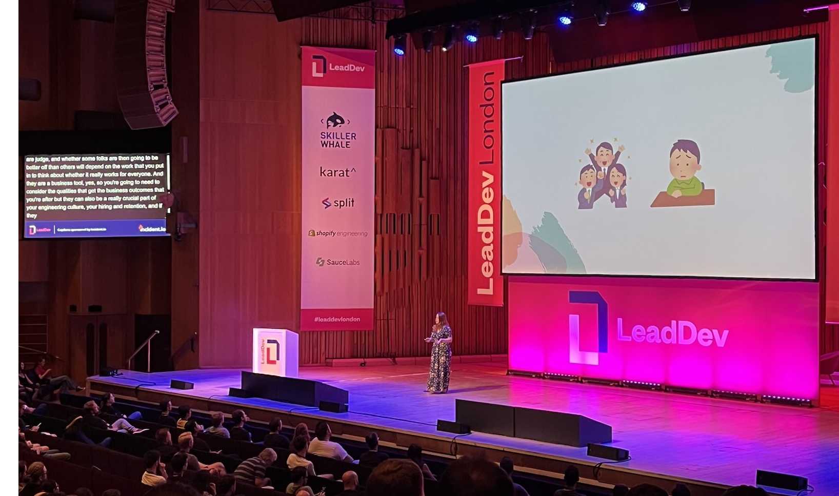 A view of me on the Lead Dev stage at the Barbican. I'm wearing a long floral dress, and am on a huge stage in front of slides with illustrations of happy and sad people on. There's lots of red and pink and Lead Dev banners around. 