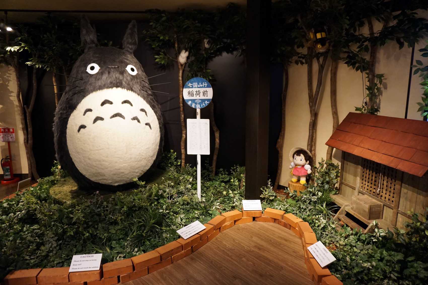 A large model of Totoro stands alongside a bus stop. To its right is a small Mei and a house model