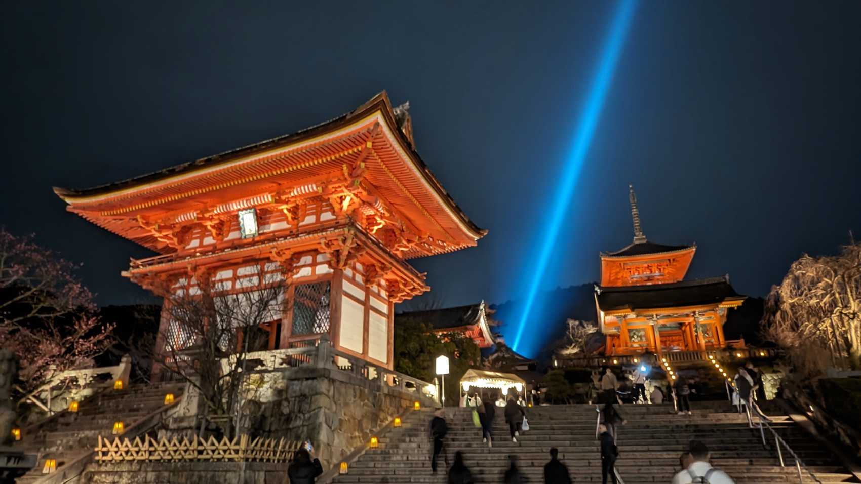 At night, a bright blue beam of light shines up at an angle between two reddy orange temple buildings from the Kiyomizudera complex