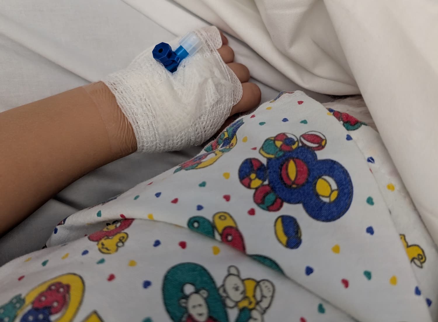 A toddler's hand, wrapped in a bandage with a cannula inserted. It's next to a colourful child's hospital gown, with numbers and bears on it.