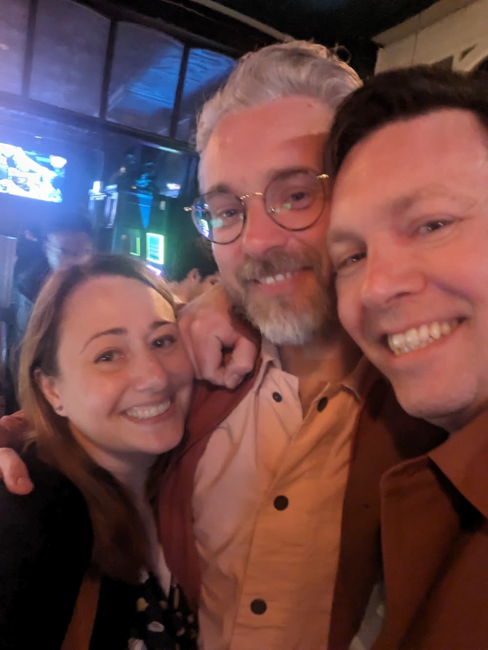 Sally, grinning at the camera, with two friends. The man in the middle has silver hair and glasses, and the man on the right has dark hair