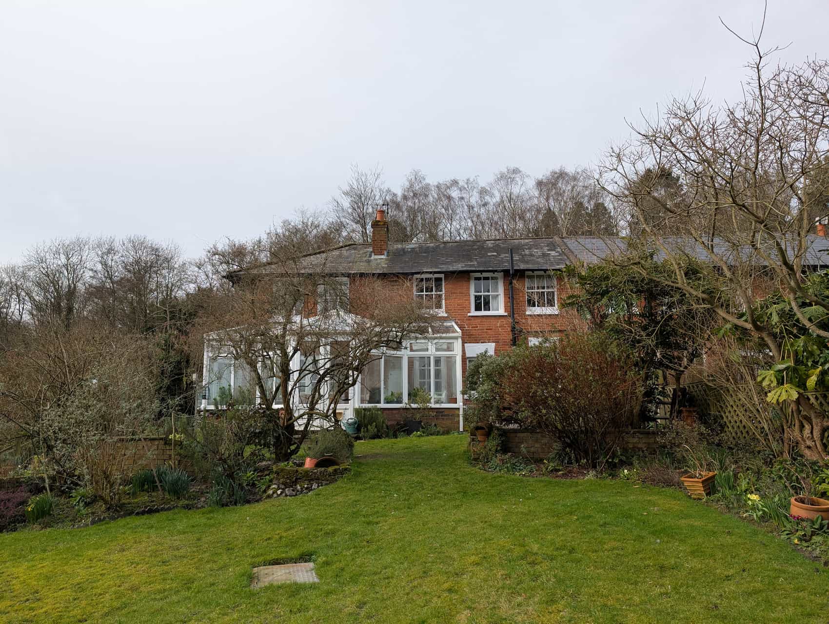 View of the back of a brick semi detached house, with a white PCV conservatory, surrounded by garden
