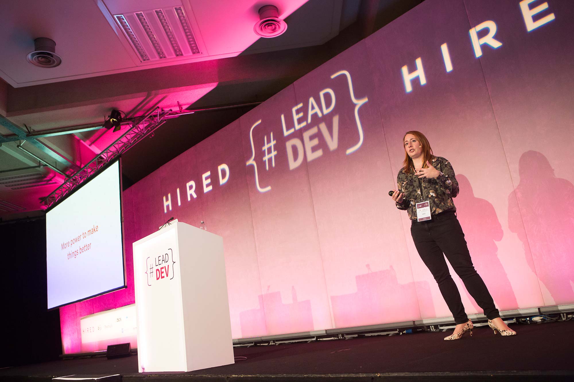 Sally standing next to a white podium on a stage with a pink backdrop, where LeadDev and Hired are displayed prominently. She's wearing a green shirt and black jeans, and holding a clicker.